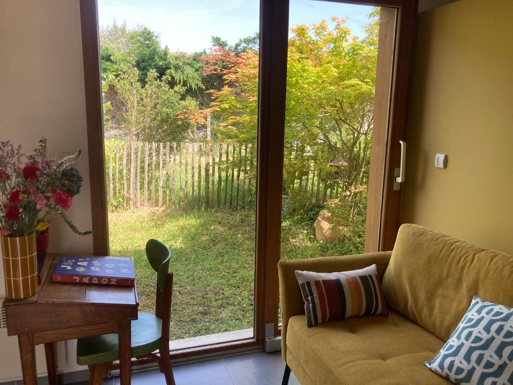 Salle de consultation avec canapé moutarde, petit secrétaire en bois et baie vitrée ouvrant sur le jardin